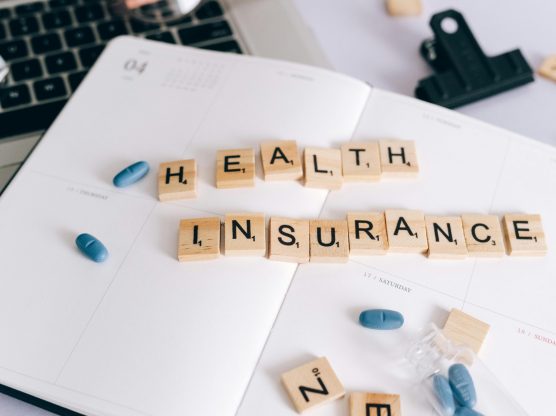 Scrabble tiles spelling 'Health Insurance' on planner with pills and laptop, symbolizing healthcare planning.